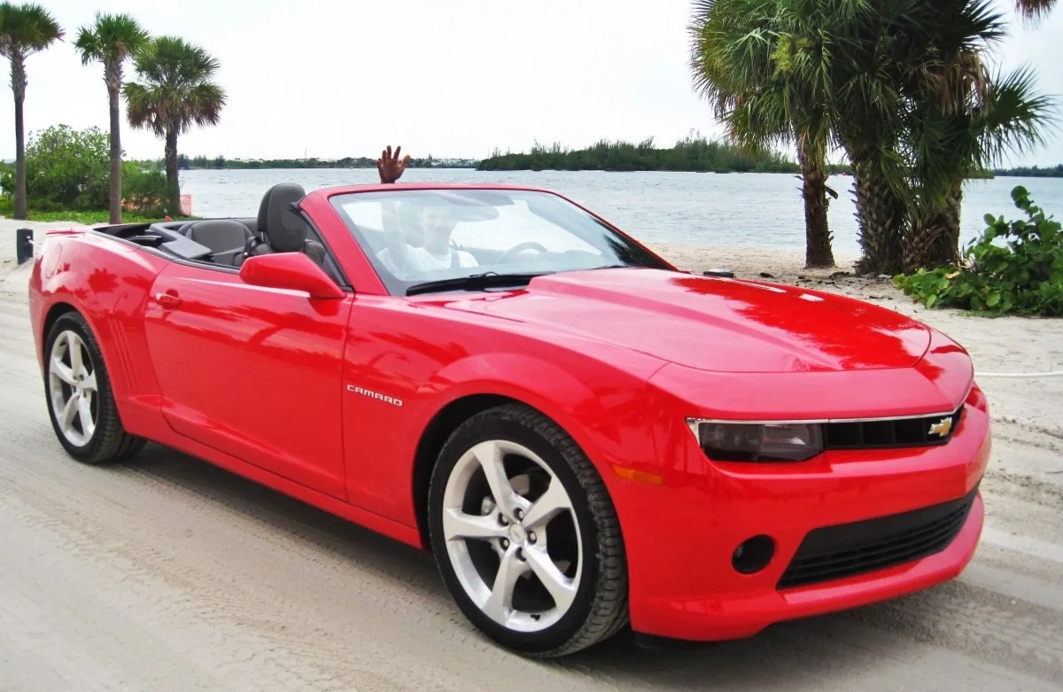 Red Chevrolet Camaro convertible parked on a sandy road with a person waving from the driver's seat.